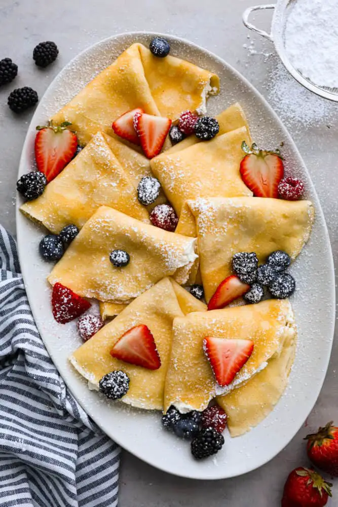 Overhead view of folded crepes on a gray platter dusted with powdered sugar and topped with fresh berries. - Perfect Crepes
