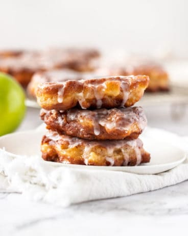 A stack of 3 apple fritters on a white plate with more in the background - White Chocolate Sauce