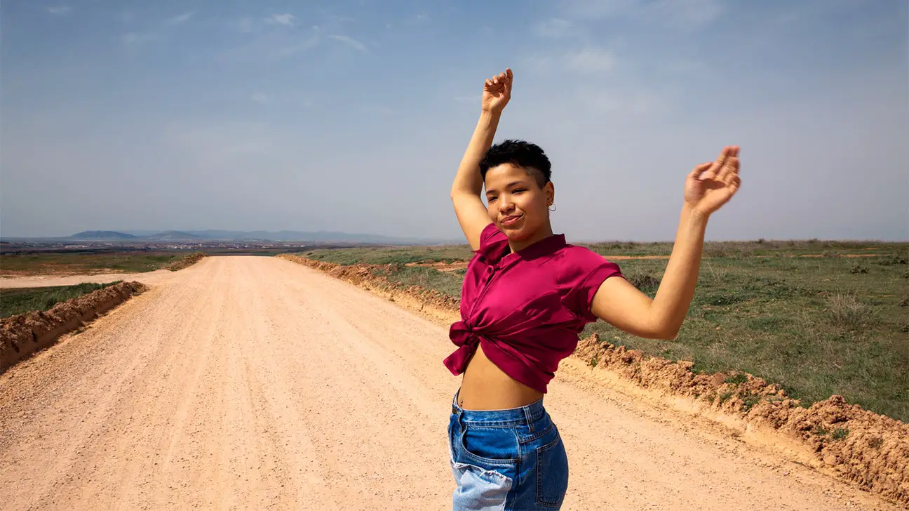 Woman with hands up, blue sky and open road in background - Quiz: What's Your 2023 Wellness Vibe?
