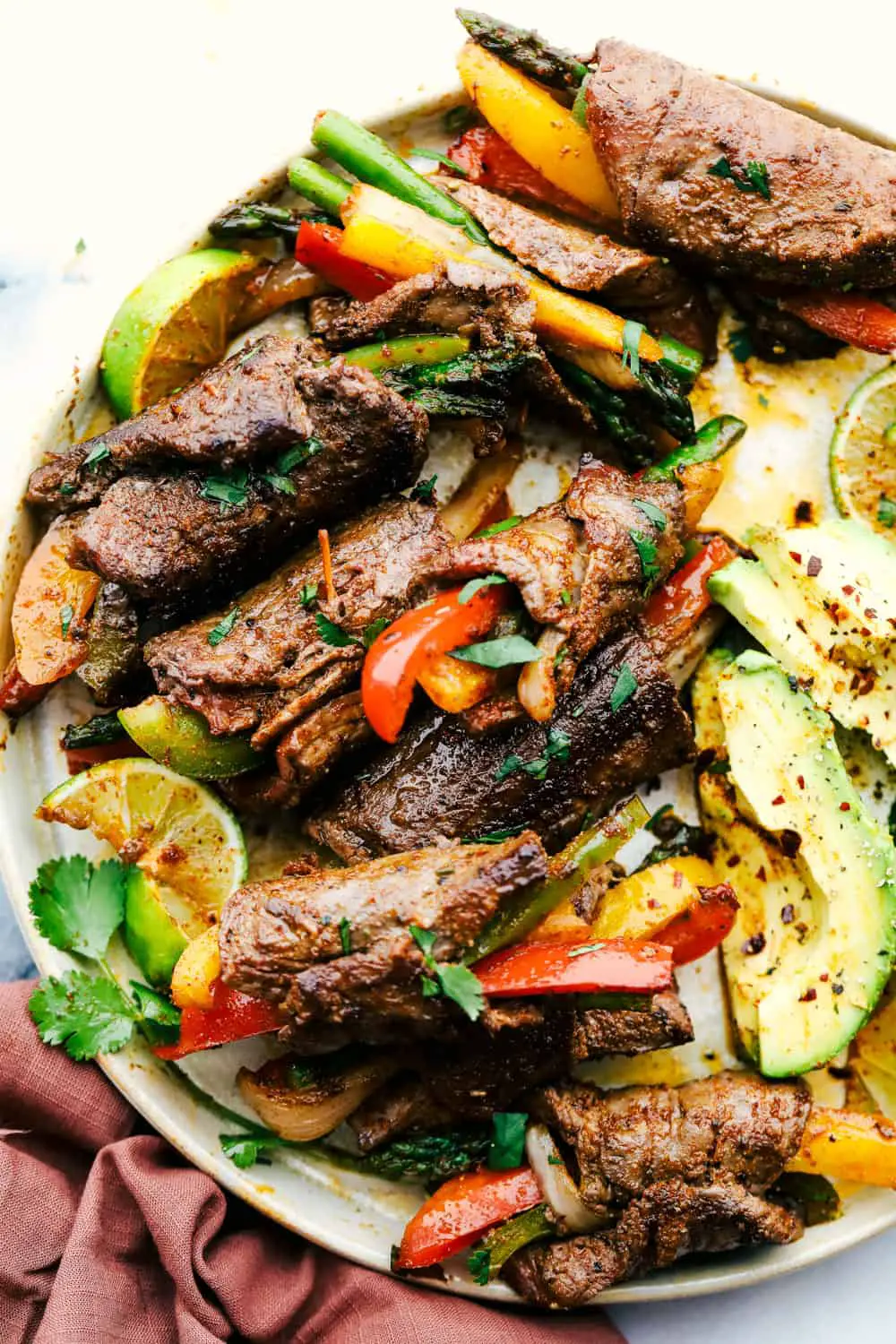 Bell peppers and asparagus wrapped up in flank steak with avocados on a plate. - Steak Fajita Roll Ups