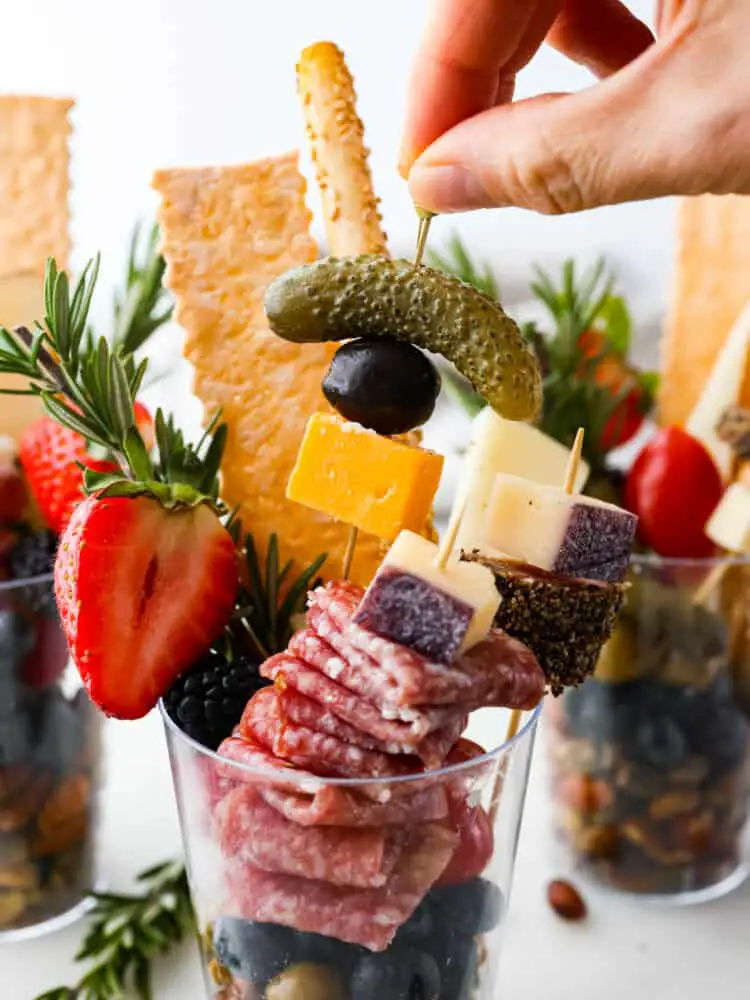 Front view of a plastic cup filled with nuts, meats, and cheeses. A skewer with a pickle, olive, and slice of cheese is being picked up. - Charcuterie Cups