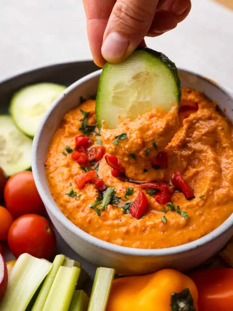 Red pepper hummus in a bowl with a cucumber being dipped in it. - Roasted Red Pepper Hummus