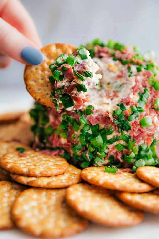 Chipped beef cheese ball with a hand dipping a cracker into it. - Easy Chipped Beef Cheese Ball