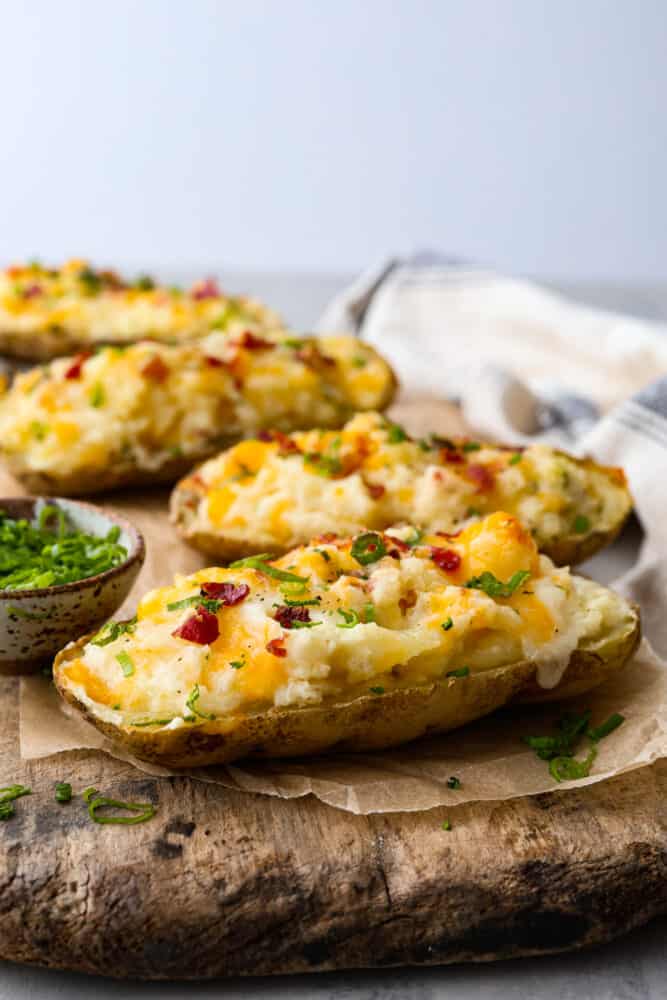 Twice-baked potatoes topped with bacon, cheese, and green onions, served on a wooden board. - Twice Baked Potatoes