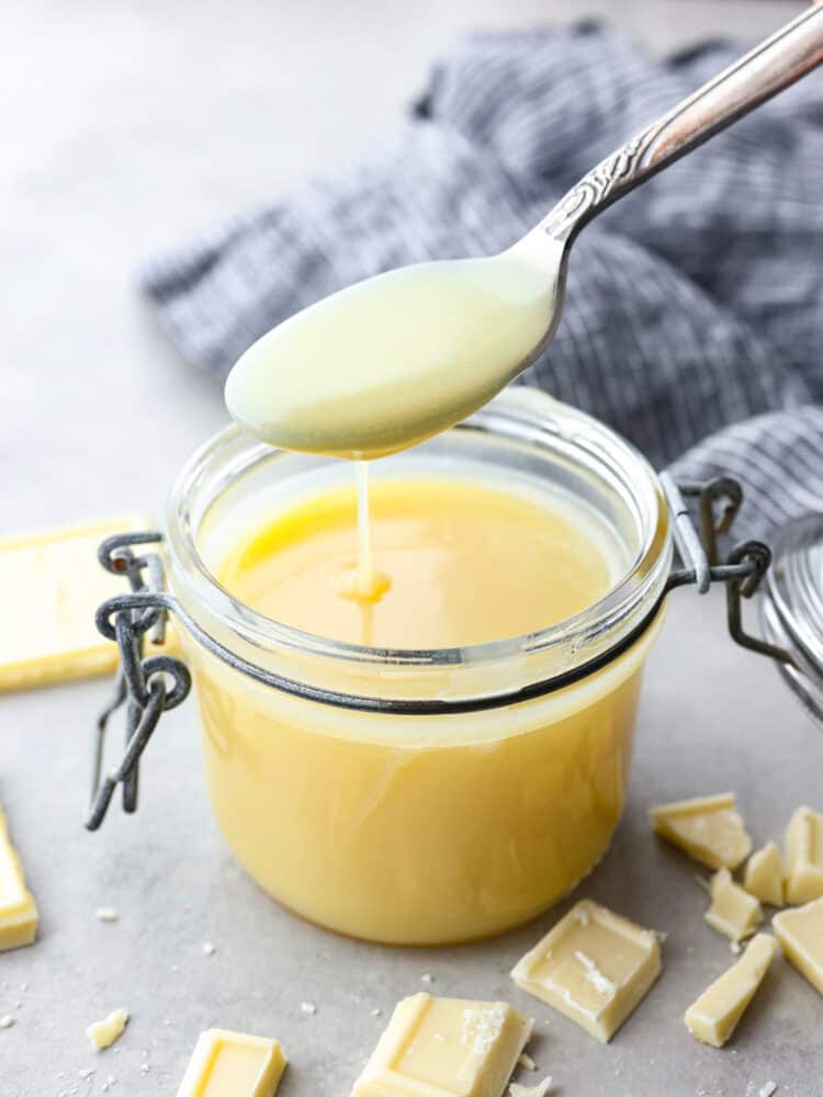 A spoon taking a scoop out of the sauce jar with white chocolate sprinkled around it. - White Chocolate Sauce